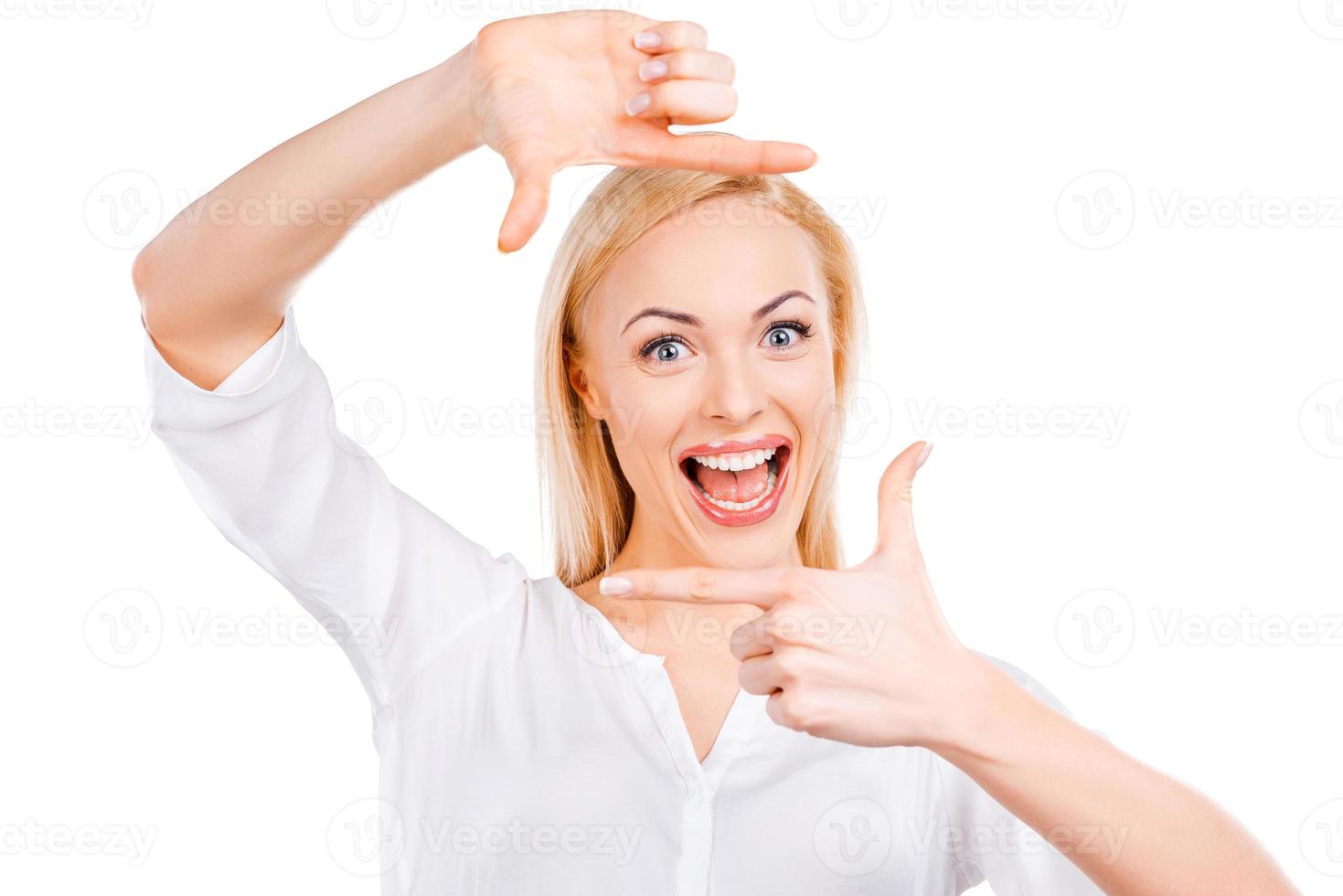 retrato de la felicidad. feliz joven mujer de pelo rubio mirando a la cámara y gesticulando marco de dedo mientras está de pie contra el fondo blanco foto