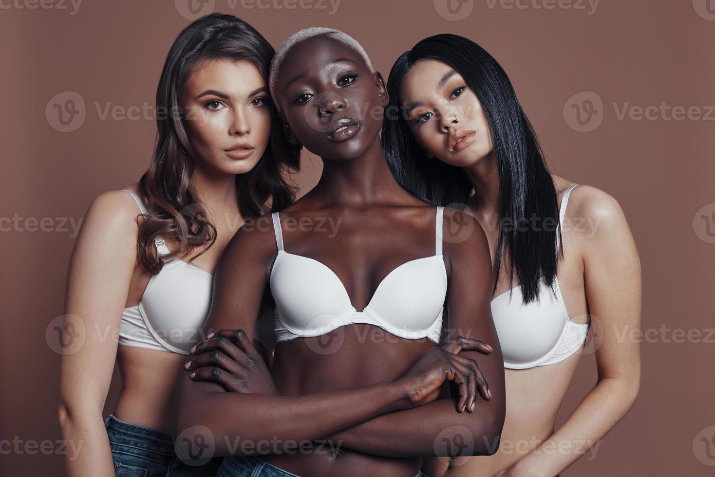 Too good to be real. Three attractive young women looking at camera while standing against brown background photo