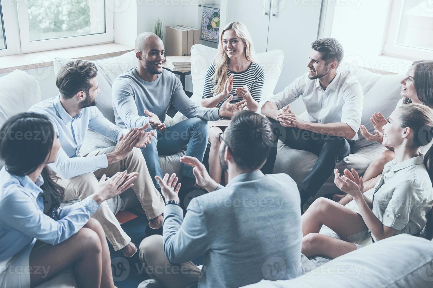 aplaudiendo sus logros. grupo de jóvenes alegres sentados en círculo y aplaudiendo mientras un hombre gesticula y sonríe foto