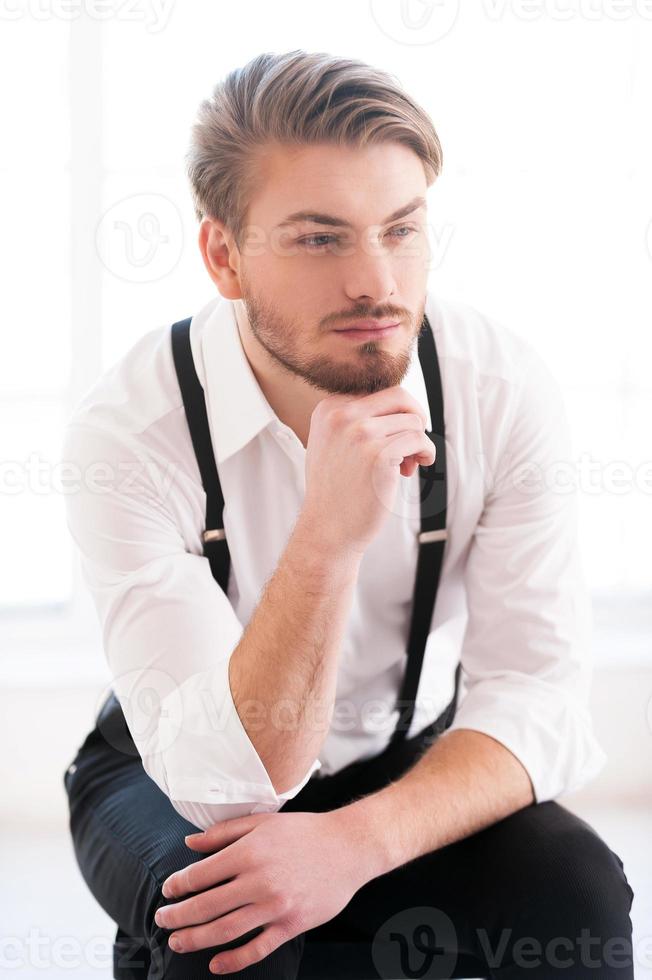 buscando inspiración. un joven pensativo con camisa y tirantes sosteniendo la mano en la barbilla y mirando hacia otro lado mientras se sienta en la silla foto