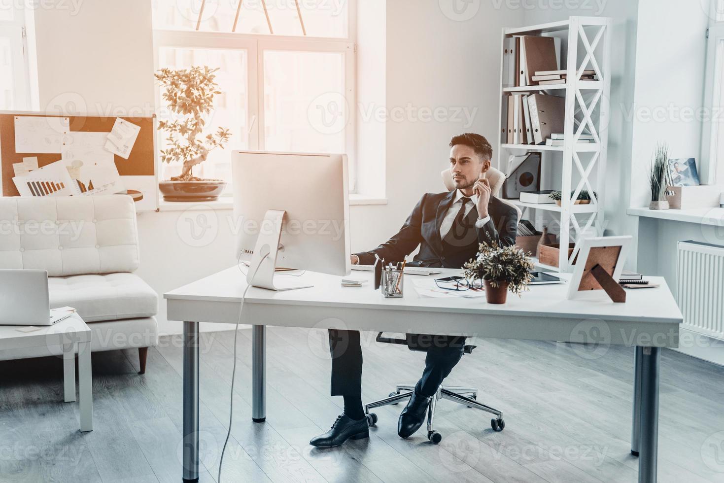 Thinking about new solutions.  Handsome young man in formalwear looking at computer monitor while sitting at the office desk photo