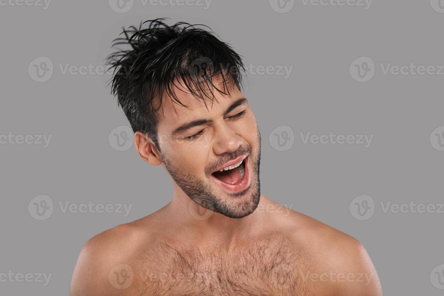 Feeling free to do anything. Good looking young man keeping mouth open and eyes closed while standing against grey background photo