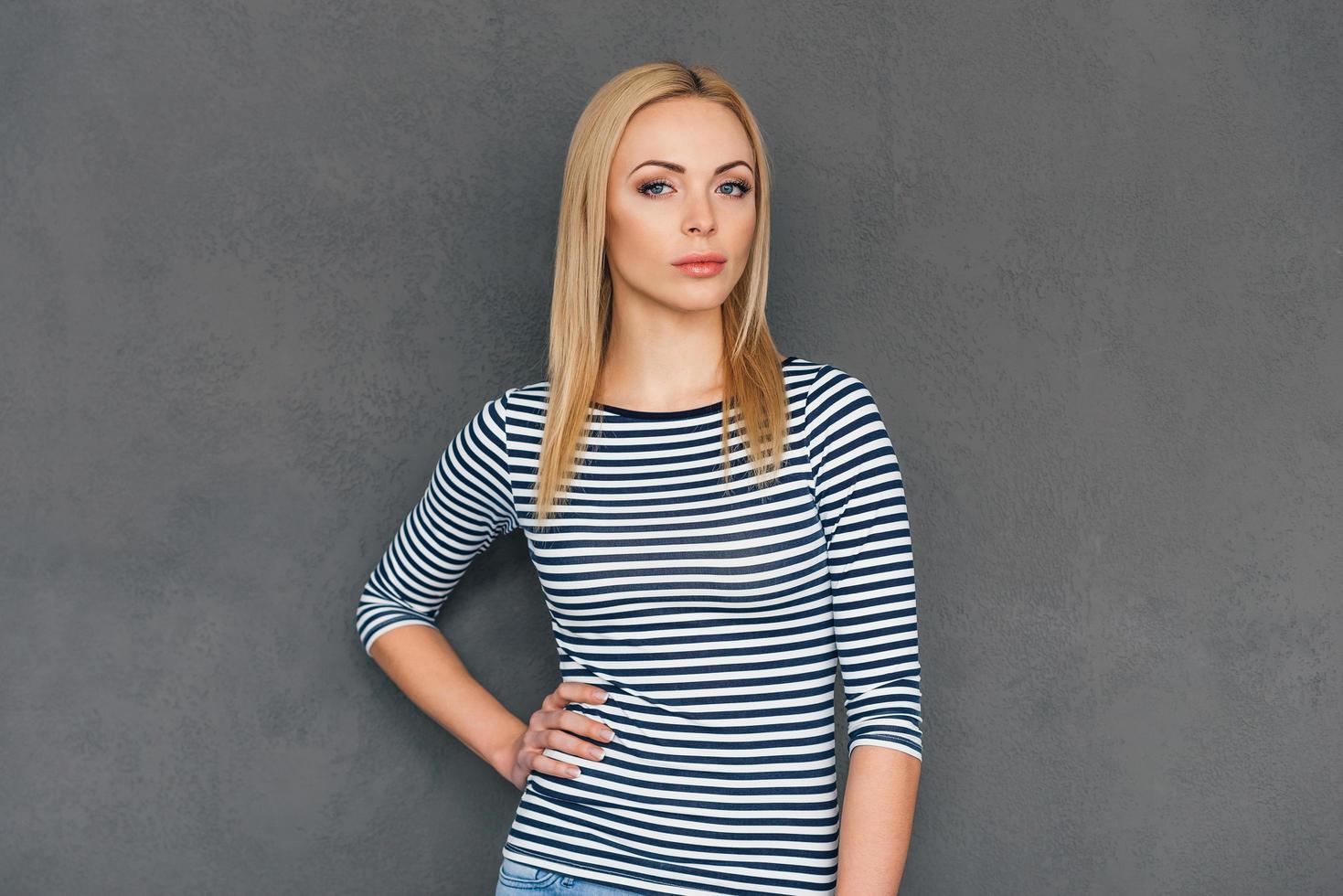 Casually beautiful. Beautiful young woman keeping hand on hip and looking at camera while standing against grey background photo