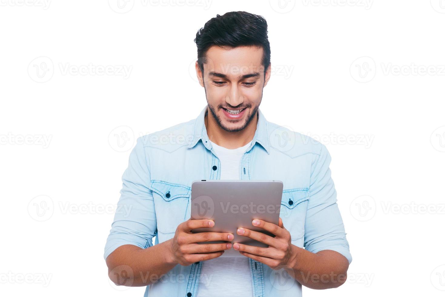 Examining his brand new gadget. Handsome young Indian man holding digital tablet and looking at it with smile while standing against white background photo