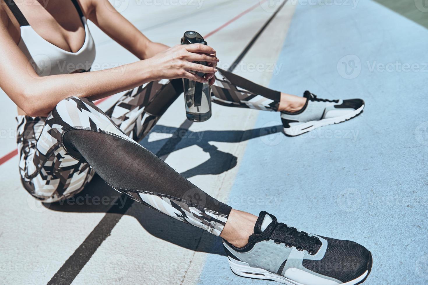 Always in a great shape. Close up top view of young woman in sports clothing holding a bottle of water while sitting on the running track outdoors photo