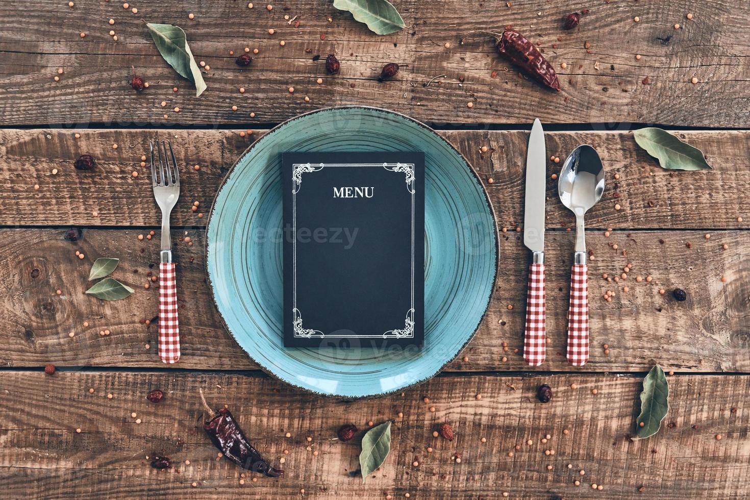 sea nuestro invitado toma en ángulo alto de plato vacío, tenedor, cuchara, cuchillo y menú cerrado sobre una mesa rústica foto