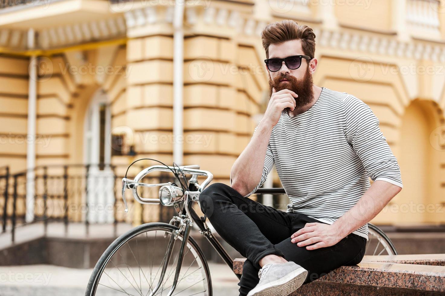 Taking quick break. Thoughtful young bearded man holding hand on chin and looking at camera while sitting near his bicycle outdoors photo