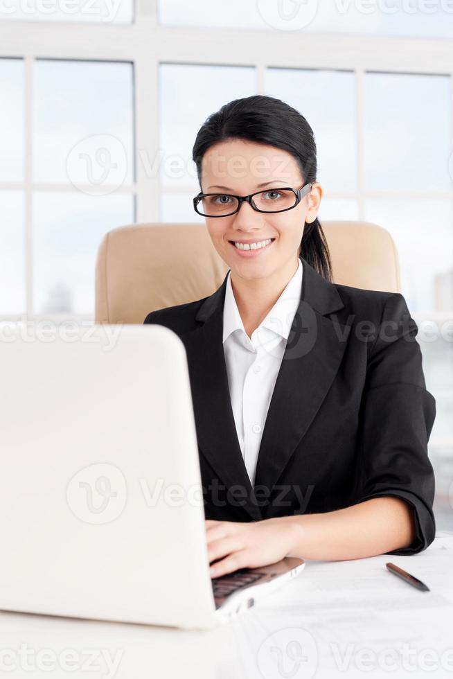 empresaria en el trabajo. mujer de negocios joven confiada que usa la computadora y sonríe mientras está sentada en su lugar de trabajo foto