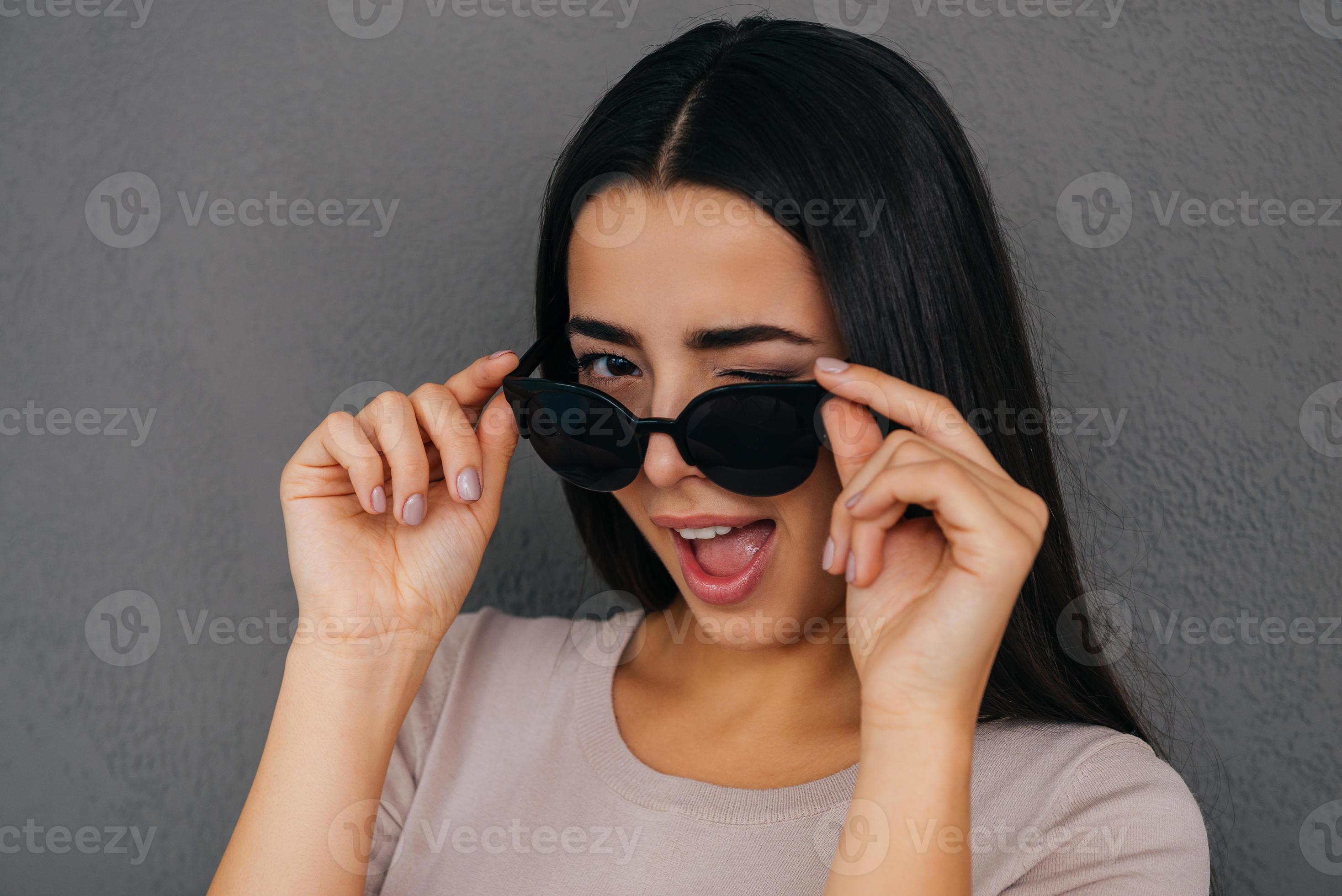 Feeling flirty. Playful young woman adjusting her sunglasses and winking to  you while standing against grey background 13503914 Stock Photo at Vecteezy