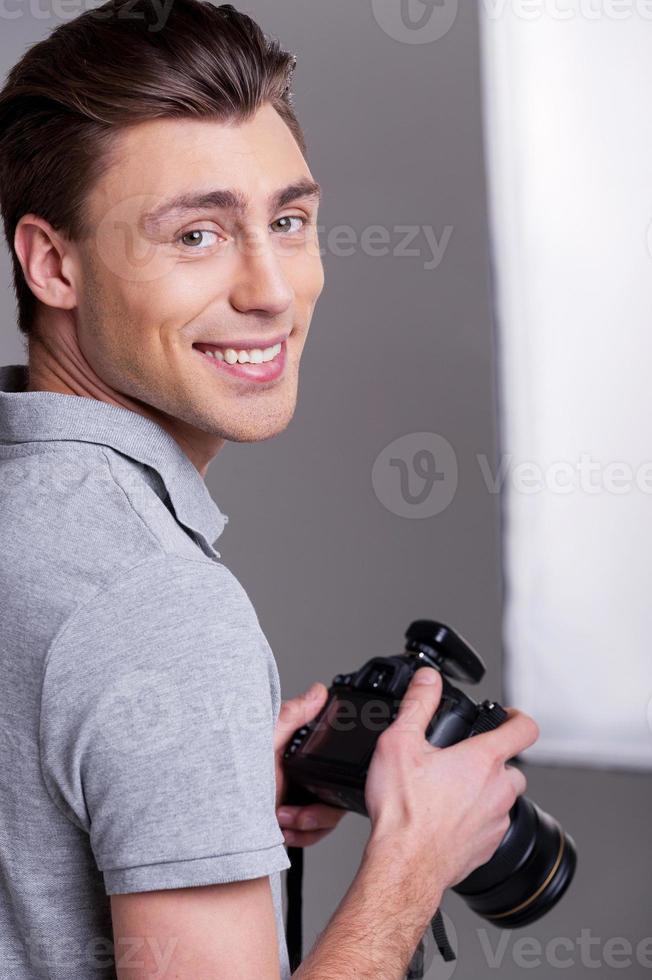 Cheerful photographer. Young man in polo shirt holding digital camera and looking over shoulder while standing in studio with lighting equipment on background photo