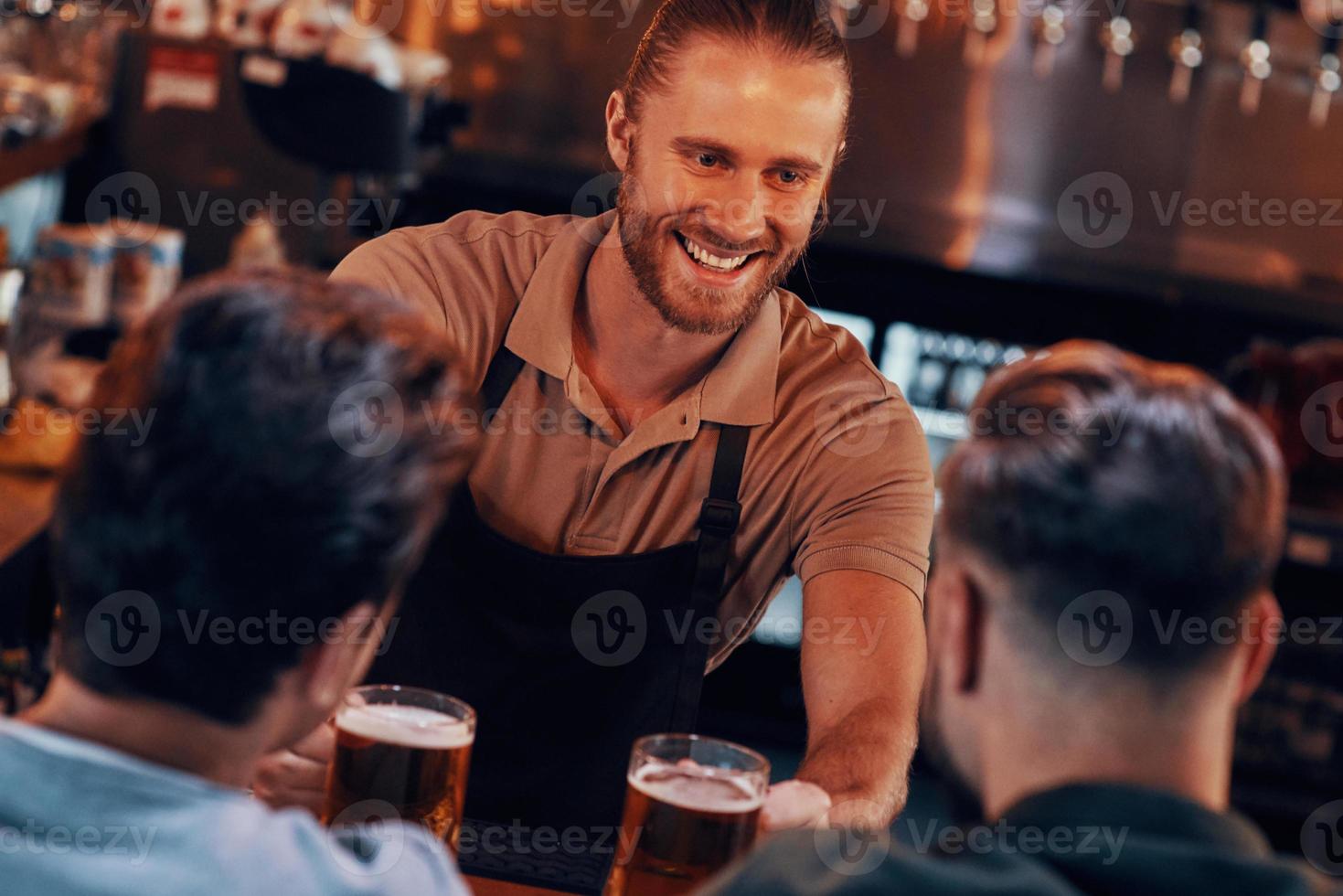 Cheerful bartender serving beer to young men while standing at the bar