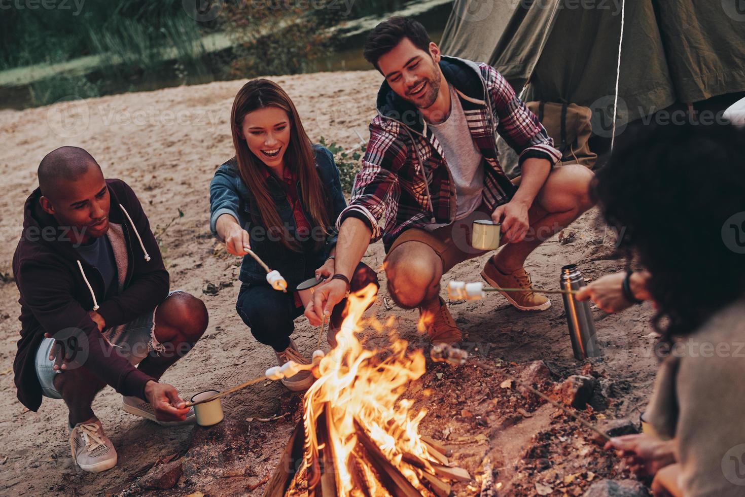 alegría de acampar. grupo de jóvenes con ropa informal asando malvaviscos sobre una fogata mientras descansan cerca del lago foto