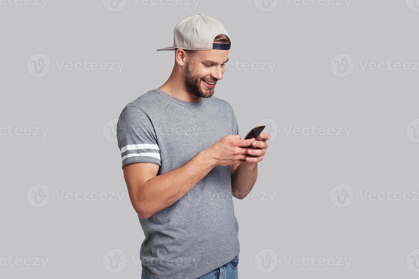 Receiving nice messages.  Handsome young man holding smart phone and looking at it while standing against grey background photo