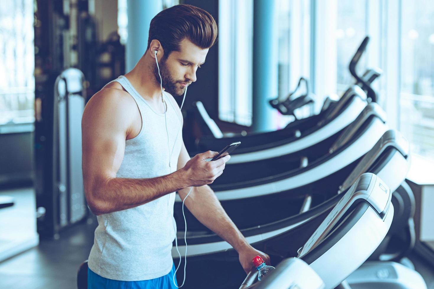 Best song for his training. Side view of young handsome man in sportswear using his smart phone while standing on treadmill at gym photo