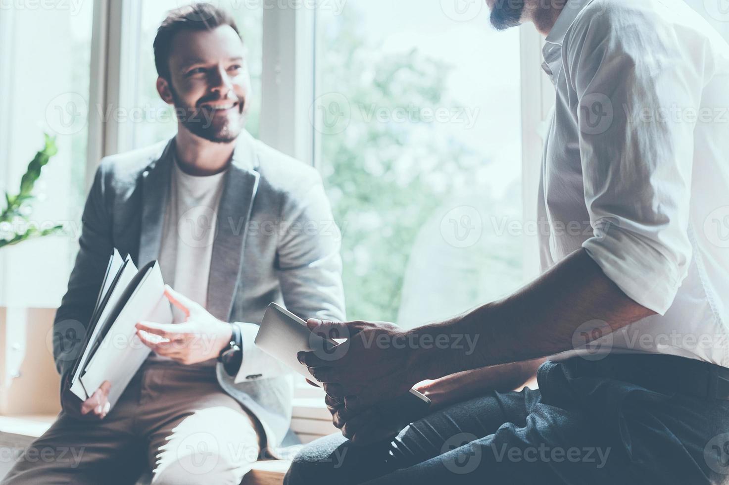 Sharing good news. Cropped image of two young businessmen in smart casual wear talking and smiling while sitting on the window sill in office photo