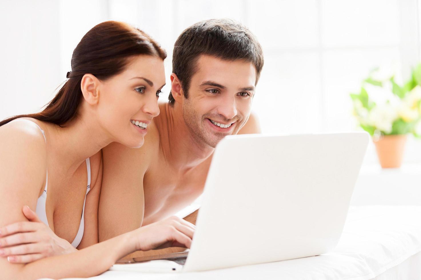 Surfing the net in bed. Cheerful young loving couple lying in bed and using computer together photo
