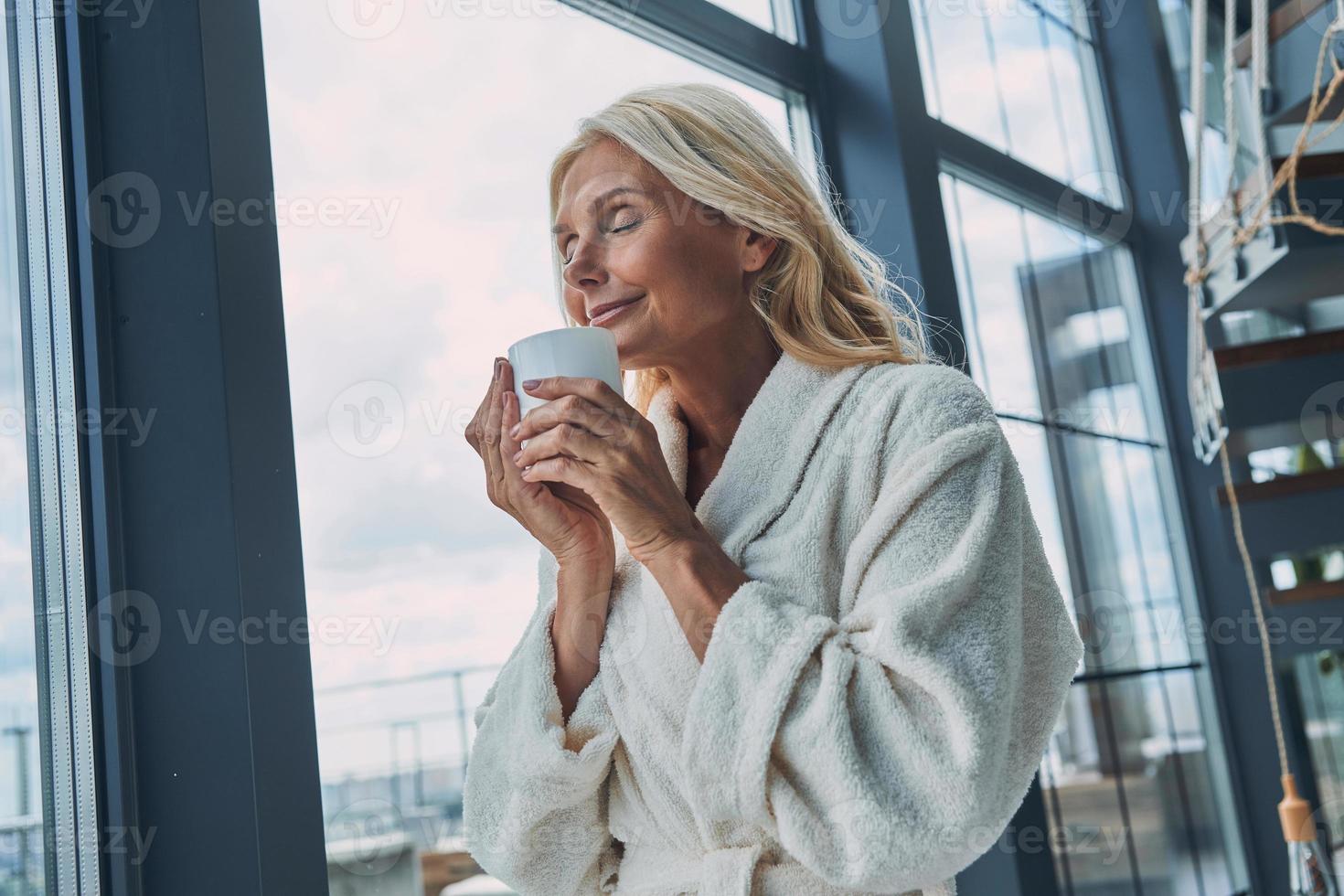 hermosa mujer madura en albornoces tomando café y manteniendo los ojos cerrados mientras está de pie cerca de la ventana en casa foto