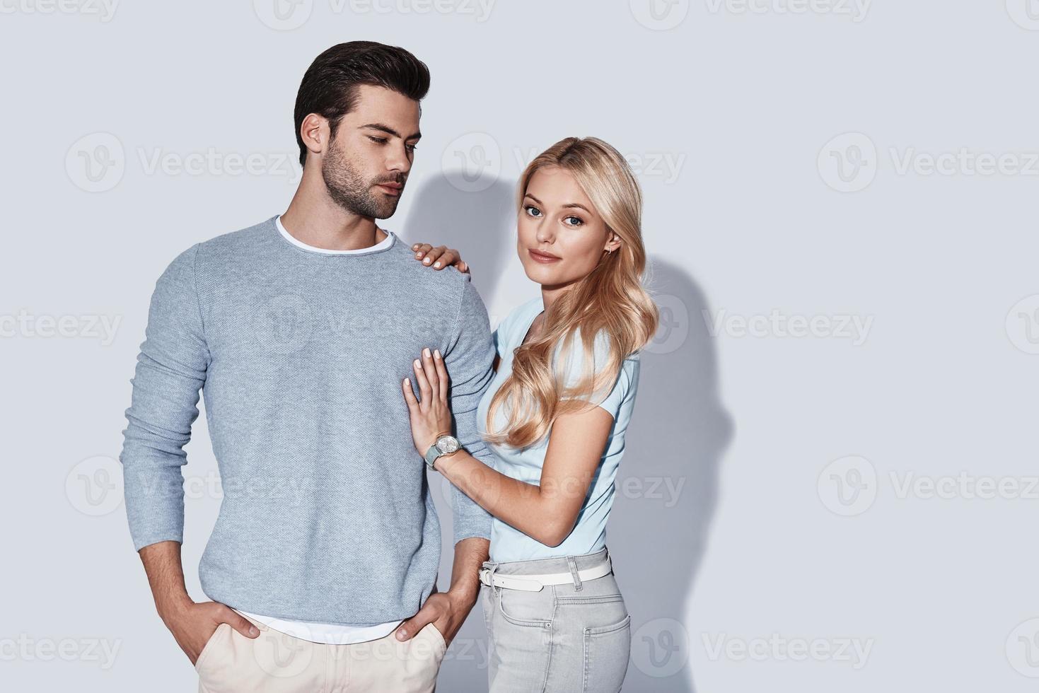 They belong together. Beautiful young couple looking at camera and smiling while standing against grey background photo