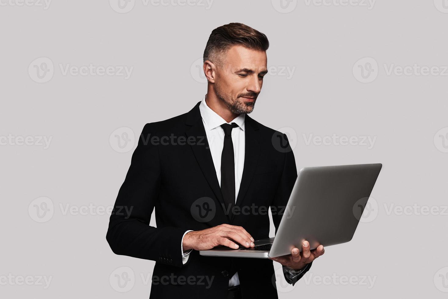 Modern technologies. Handsome young man in full suit using laptop and smiling while standing against grey background photo