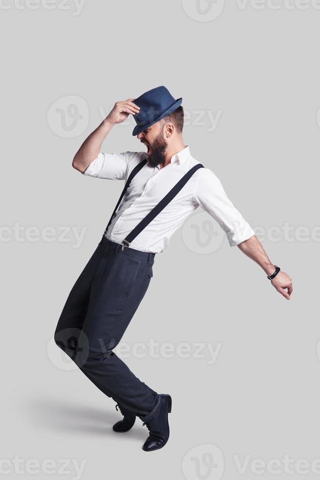 Perfecto en todo sentido. toda la longitud de un apuesto joven con tirantes ajustando su sombrero y haciendo una mueca mientras baila contra un fondo gris foto