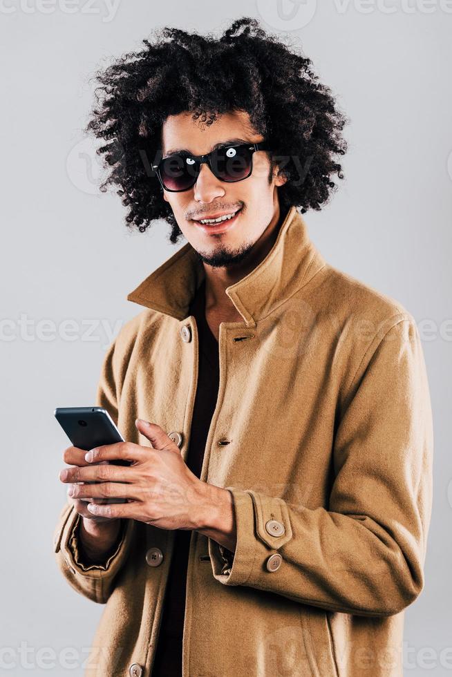 Message from friend.Cheerful young African man in sunglasses looking at camera with smile and holding smartphone while standing against grey background photo