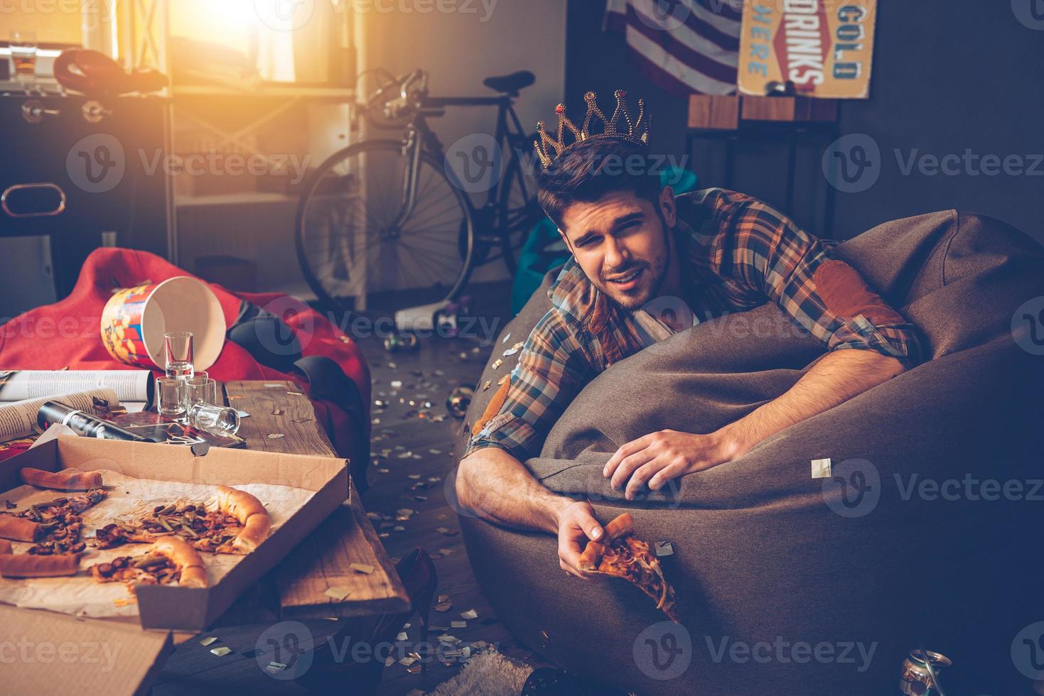 King of the party. Young handsome man in plastic crown lying on bean bag with pizza slice in his hand in messy room after party photo