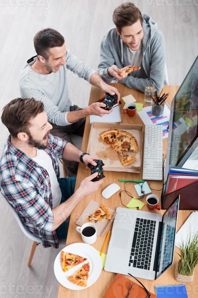 Game time. Top view of three young men playing computer games and eating pizza while sitting at the desk photo