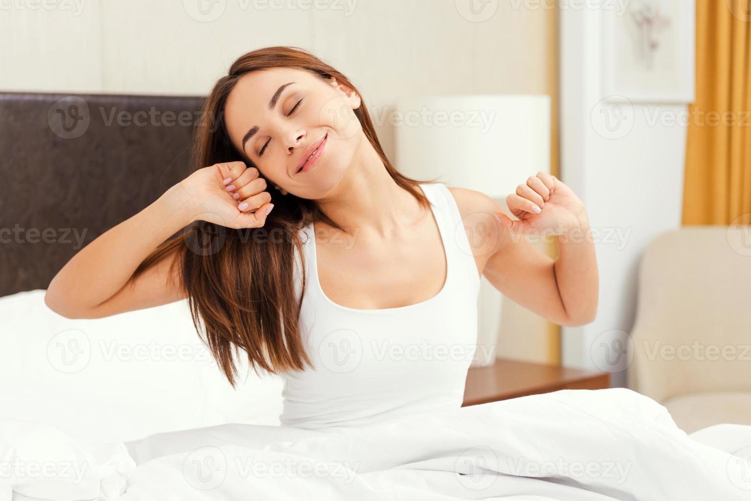 Morning stretching. Beautiful young smiling woman sitting in bed and stretching out photo