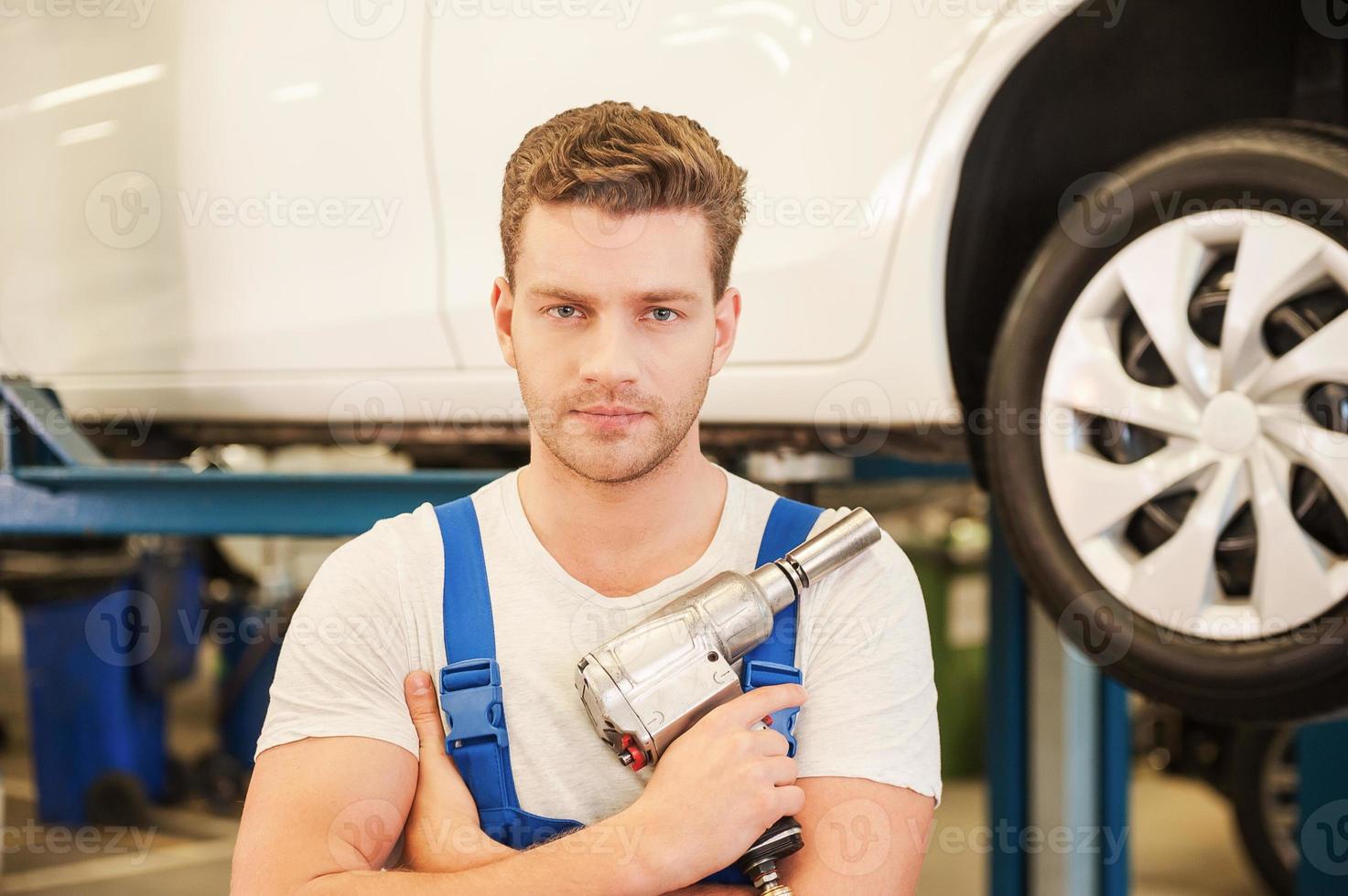 Confident mechanic. Confident young man holding a wrench and looking at camera while standing in workshop with car in the background photo