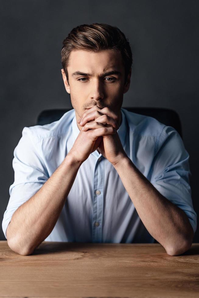 Searching for the right solution. Handsome young man keeping hands clasped and looking thoughtful while sitting against black background photo