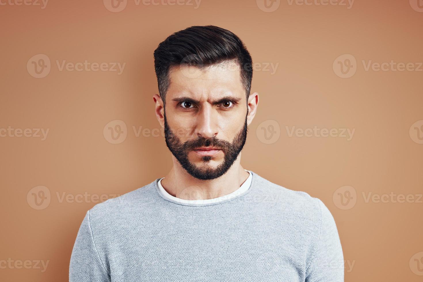 Frowning young man in casual wear looking at camera while standing against brown background photo