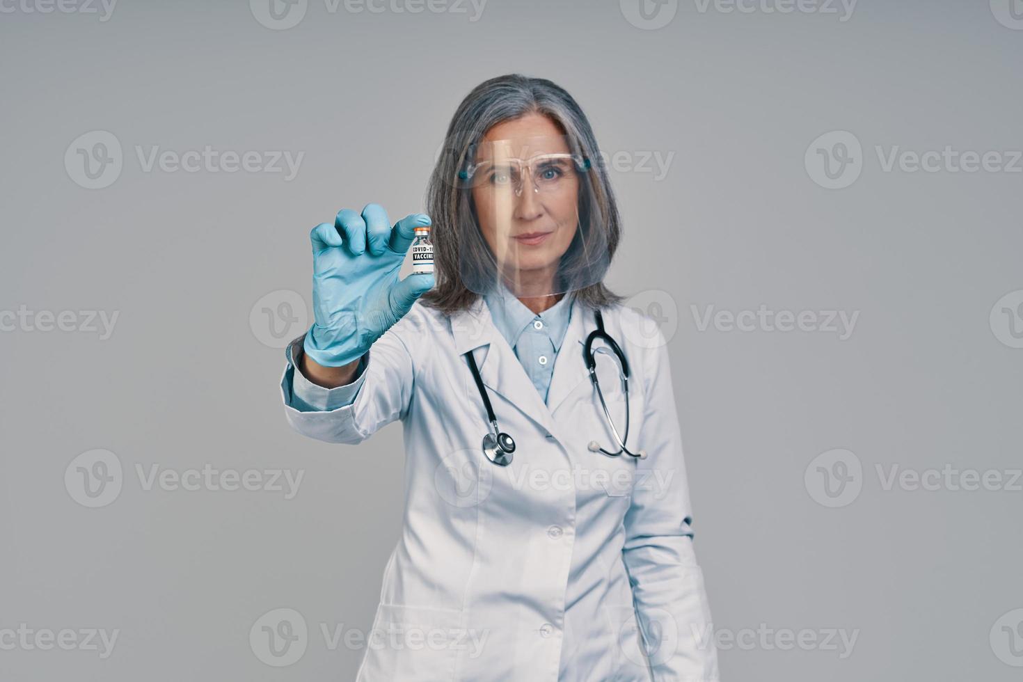 Mature beautiful female doctor in face shield holding a bottle with vaccine photo