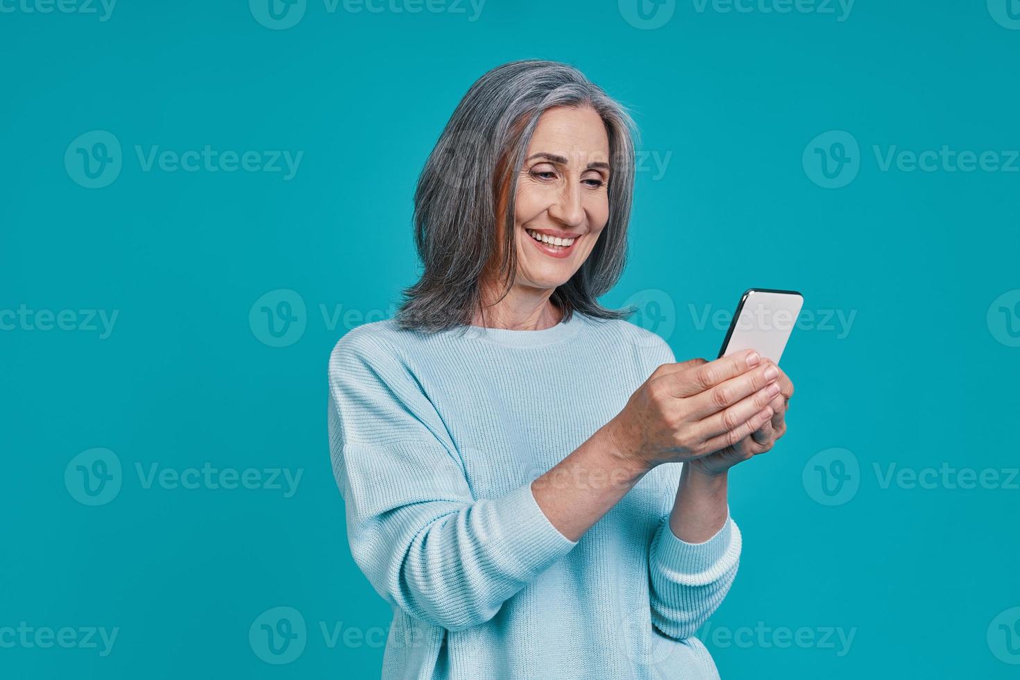 Mature beautiful woman using smart phone and smiling while standing against blue background photo