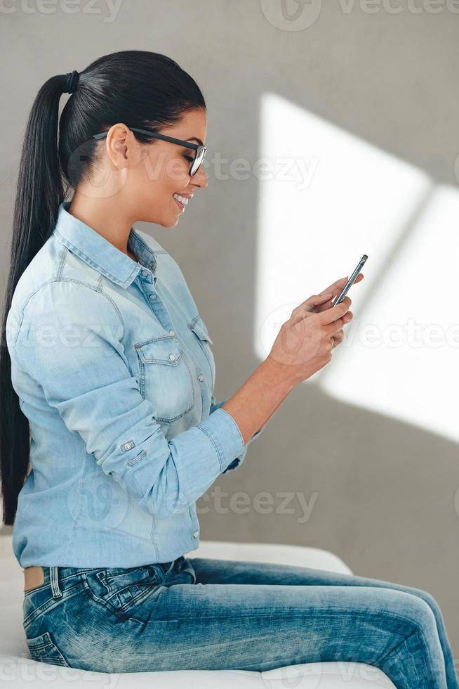 Great message from friend. Side view of beautiful young cheerful woman in glasses using her smart phone with smile while sitting on the couch against grey background photo