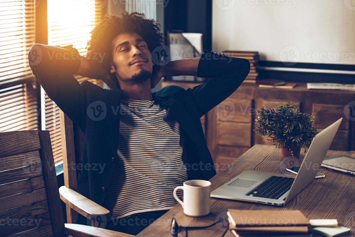 Satisfied with his work. Young handsome African man keeping hands behind his head and looking satisfied while sitting at his working place photo