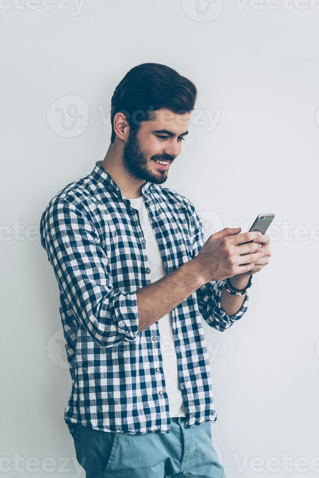 Texting to friend. Handsome young man holding smart phone and looking at it with smile photo