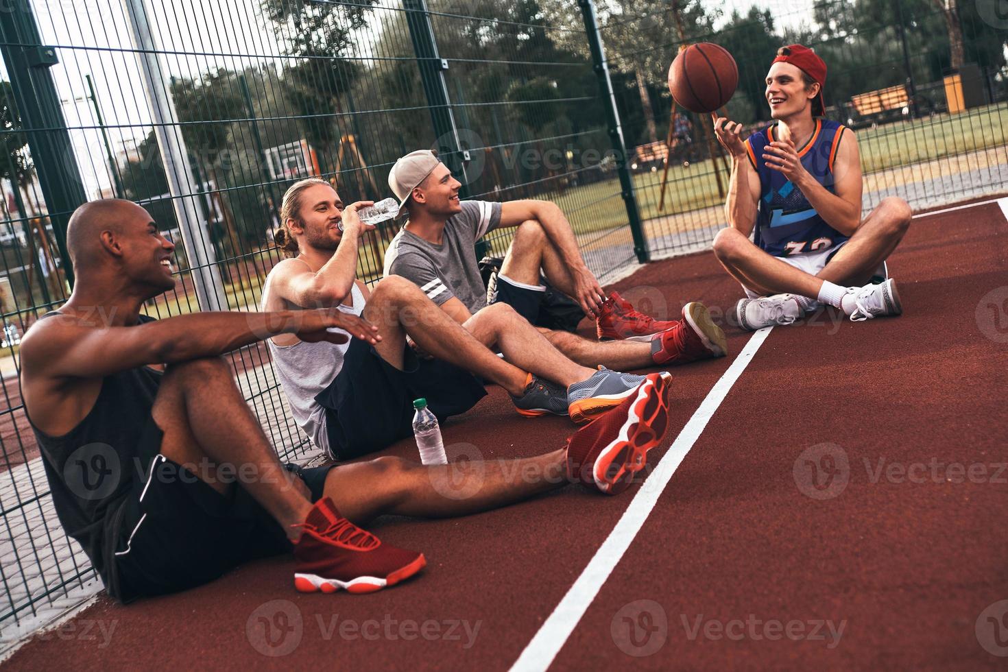 Sharing same hobbies. Group of young men in sports clothing smiling while sitting on the basketball field outdoors photo