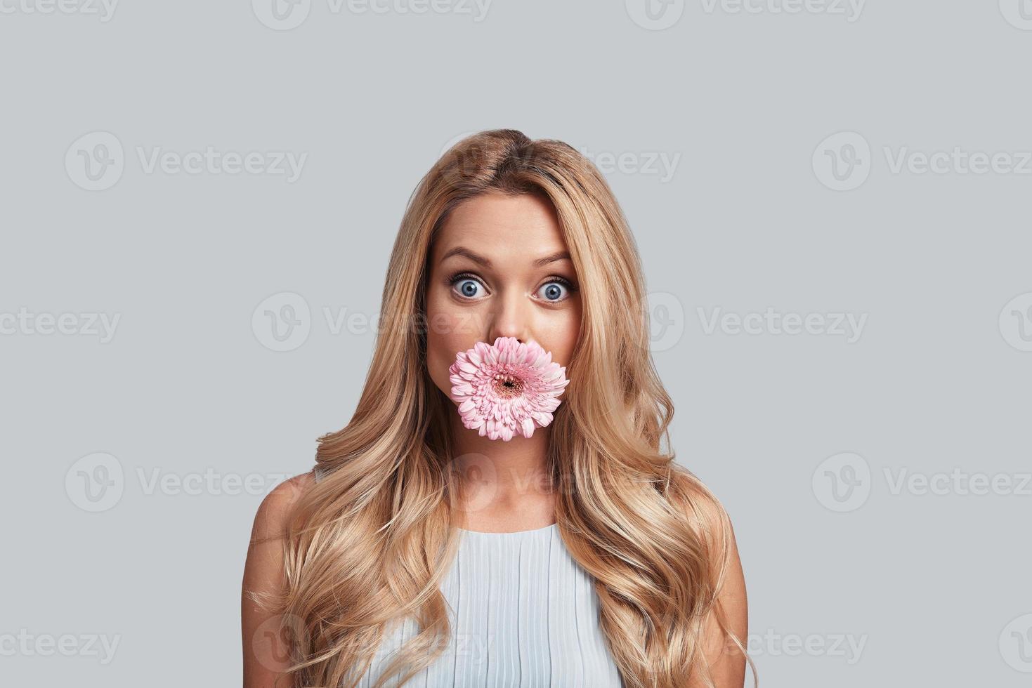 Beautiful like flower. Attractive young woman with flower covering mouth staring at camera while standing against grey background photo