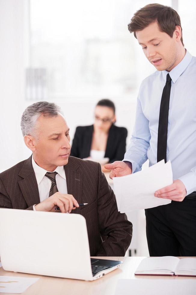 I need an advice. Two confident business people discussing something while on of them pointing paper with woman working on background photo