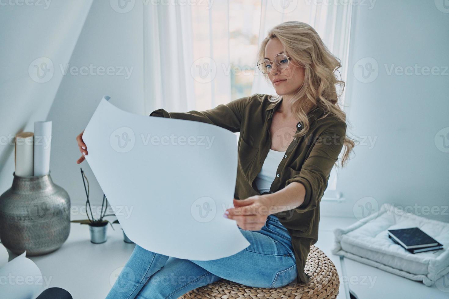 Attractive young woman exmining blueprint while sitting on padded stool at home photo