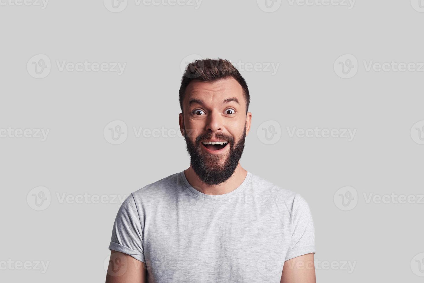 Good news. Shocked young man looking at camera and smiling while standing against grey background photo