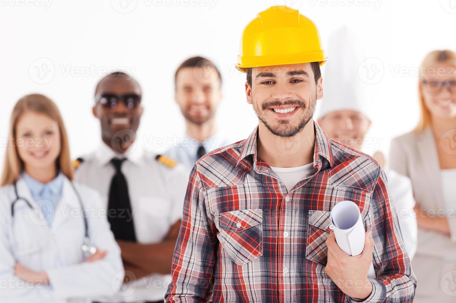 When I grow up I will be an engineer. Confident young man in hardhat holding blueprint and smiling while group of people in different professions standing in the background photo