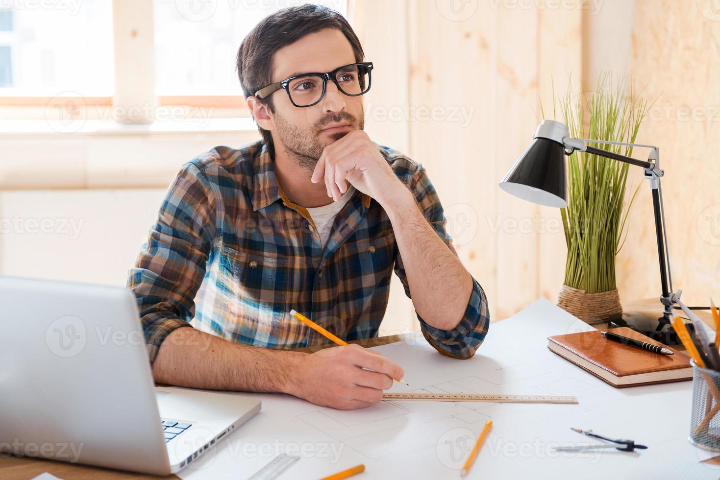 Waiting for inspiration. Thoughtful young man holding hand on chin and looking away while sitting at his working place photo