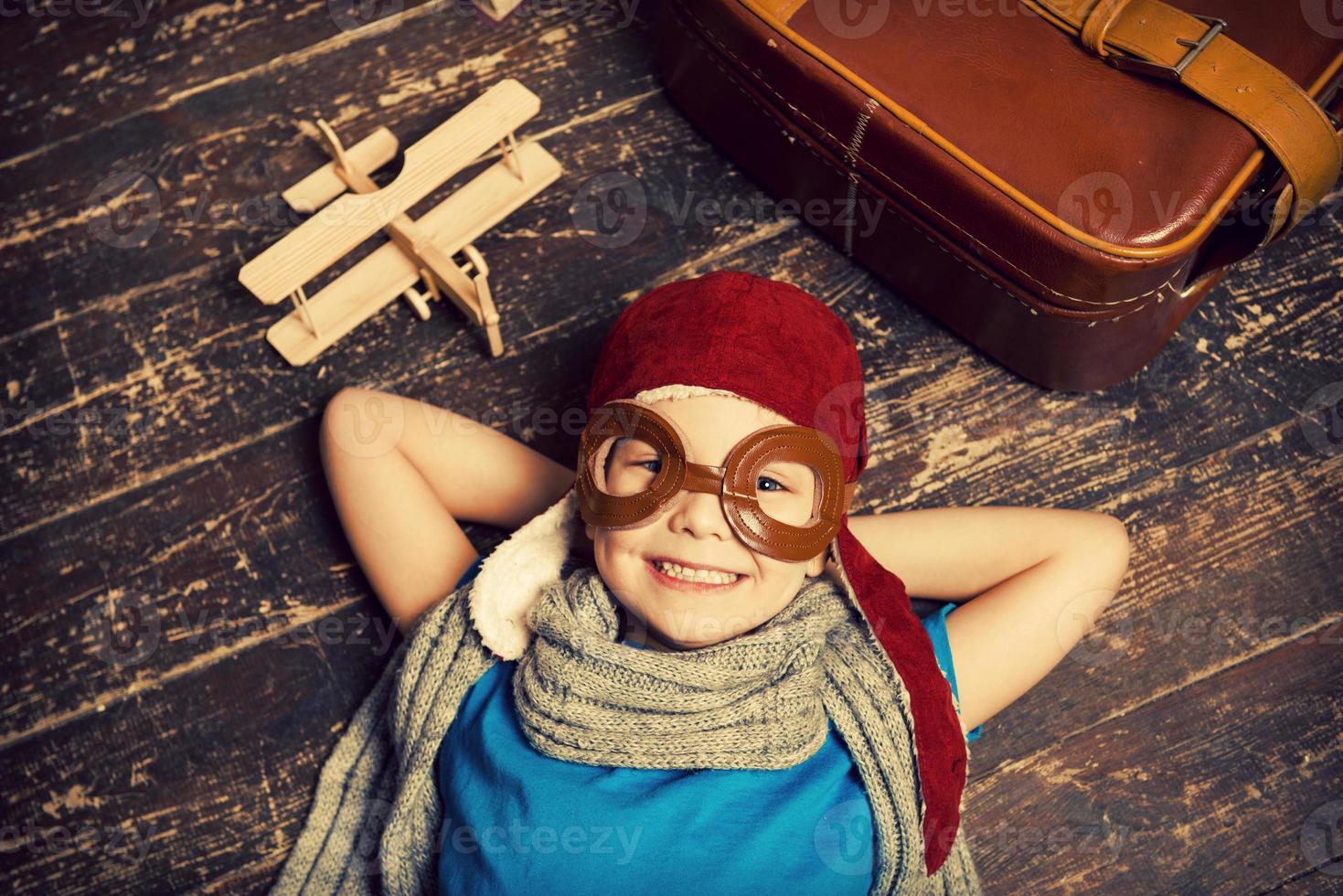 Dreaming of a big sky. Top view of happy little boy in pilot headwear and eyeglasses lying on the hardwood floor and smiling while wooden planer and briefcase laying near him photo