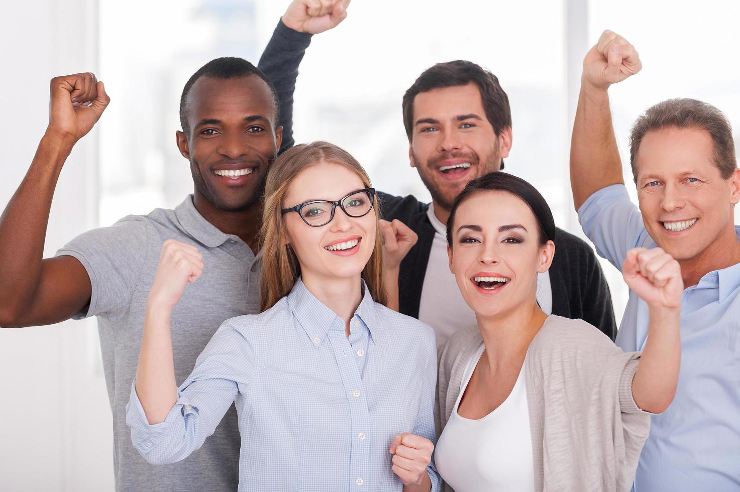 Happy business team. Group of cheerful business people in casual wear standing close to each other and keeping arms raised photo