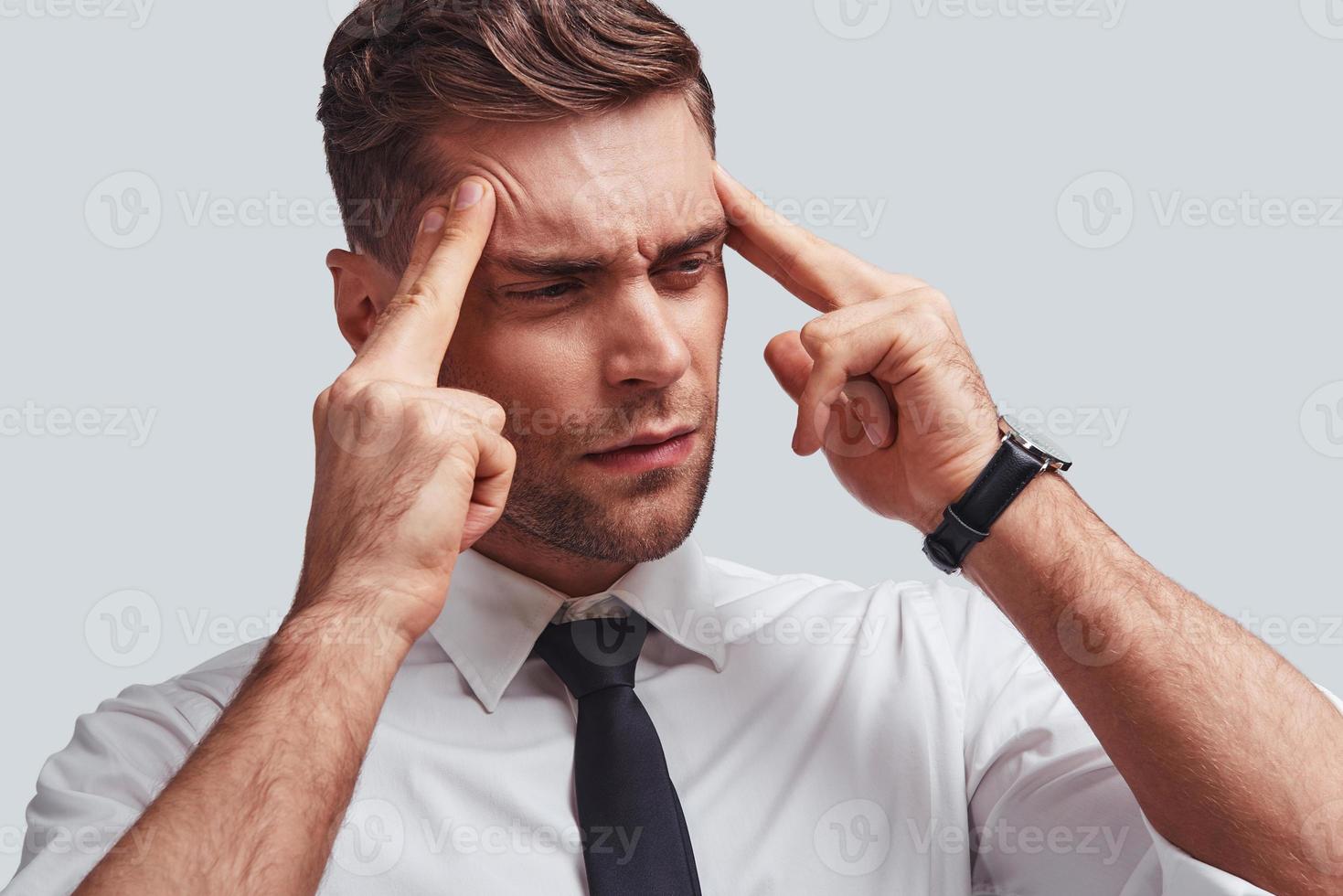 Terrible pain. Frustrated young man in full suit touching his head with hands while standing against grey background photo