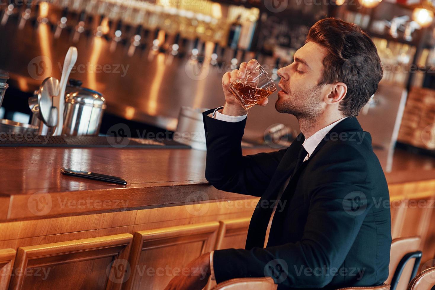 Handsome young man in full suit having alcohol drink while sitting at the bar counter in restaurant photo