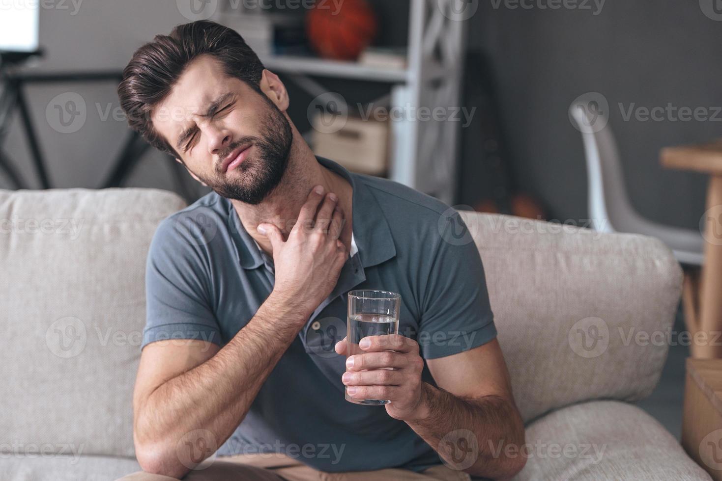 Terrible pain in his throat. Frustrated handsome young man touching his neck and holding a glass of water while sitting on the couch at home photo