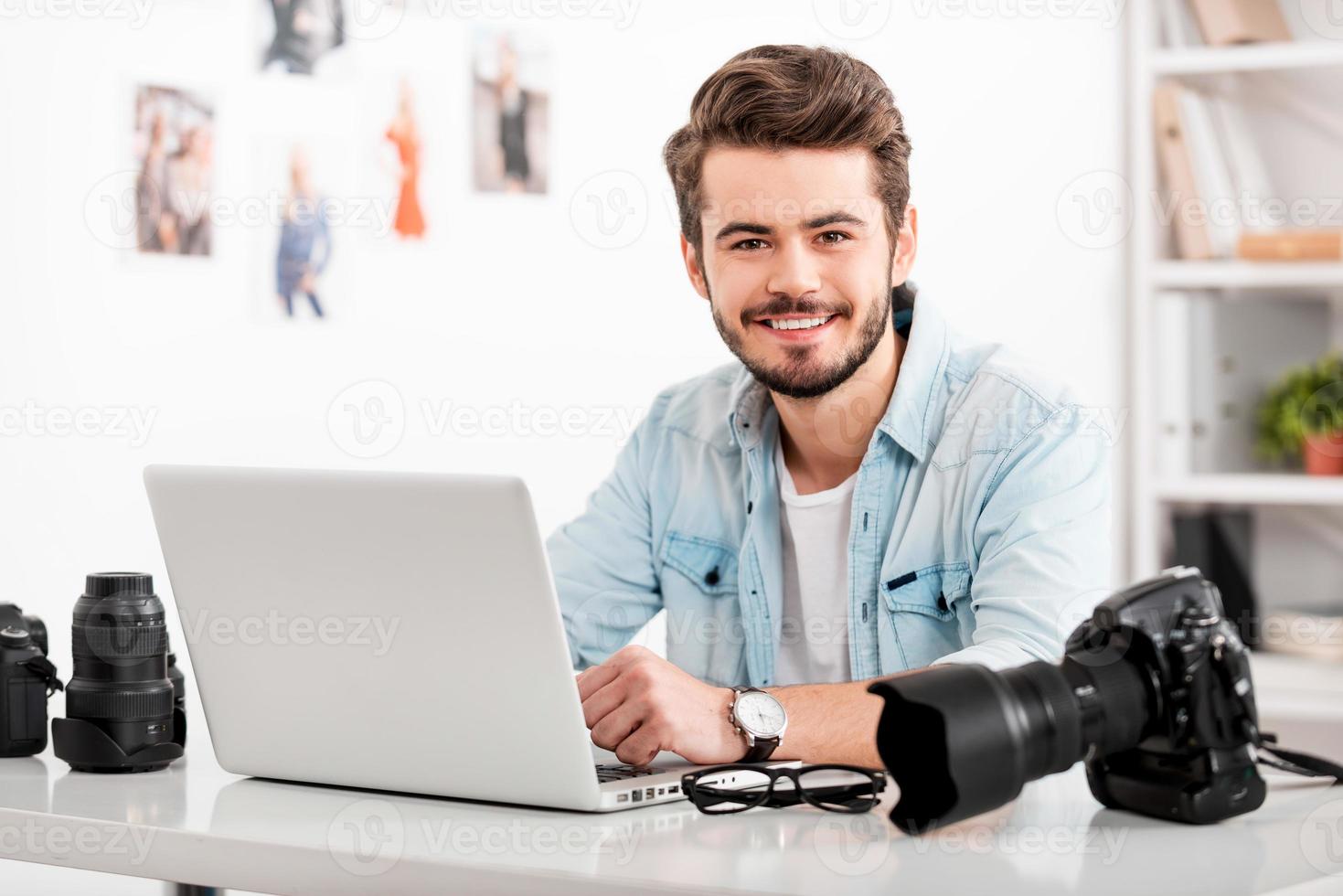 fotógrafo joven y creativo. un joven sonriente que trabaja en una laptop y mira la cámara mientras se sienta en su lugar de trabajo foto