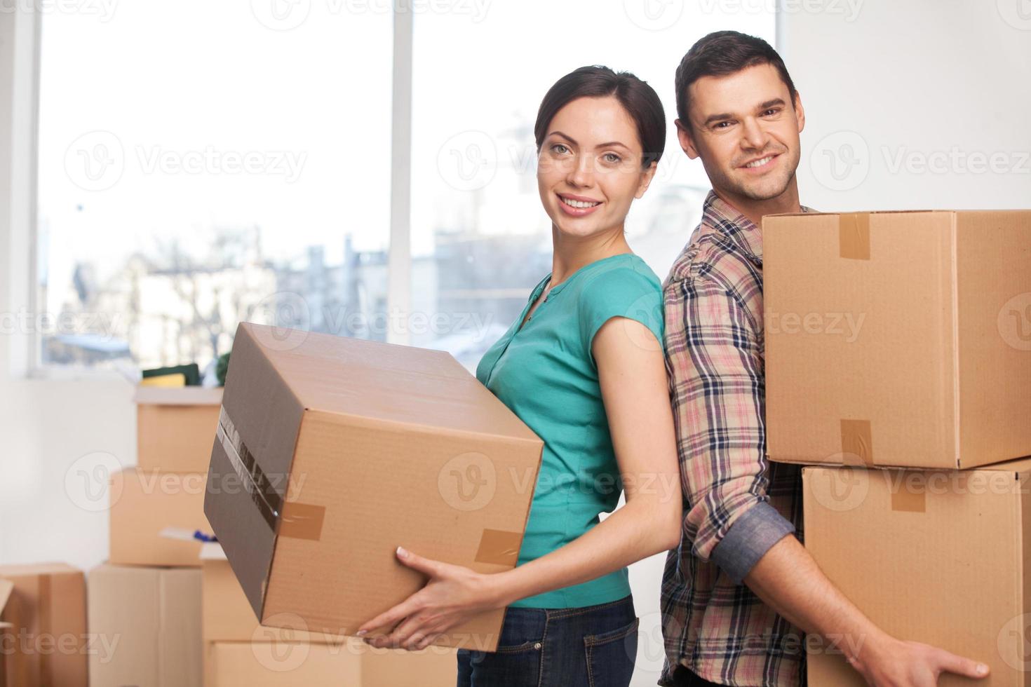 Moving to a new apartment. Beautiful young couple standing close to each other and smiling at camera while holding cardboard boxes photo